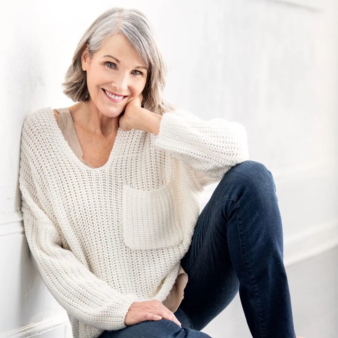 Smiling woman with shoulder-length gray hair, wearing a white knit sweater and blue jeans, sitting with one leg up. She leans against a white wall, resting her hand on her knee. The room is softly lit, creating a warm and relaxed atmosphere.